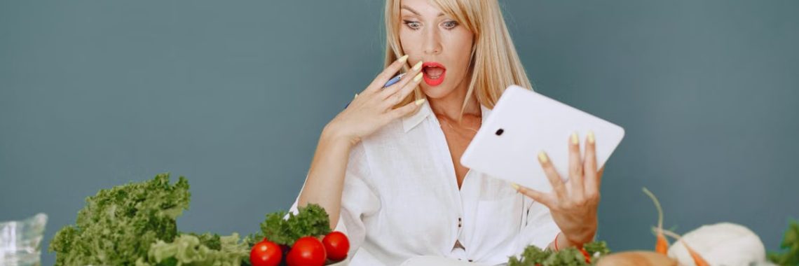 A surprised woman looking at a tablet surrounded by fresh vegetables and fruits, promoting healthy eating.