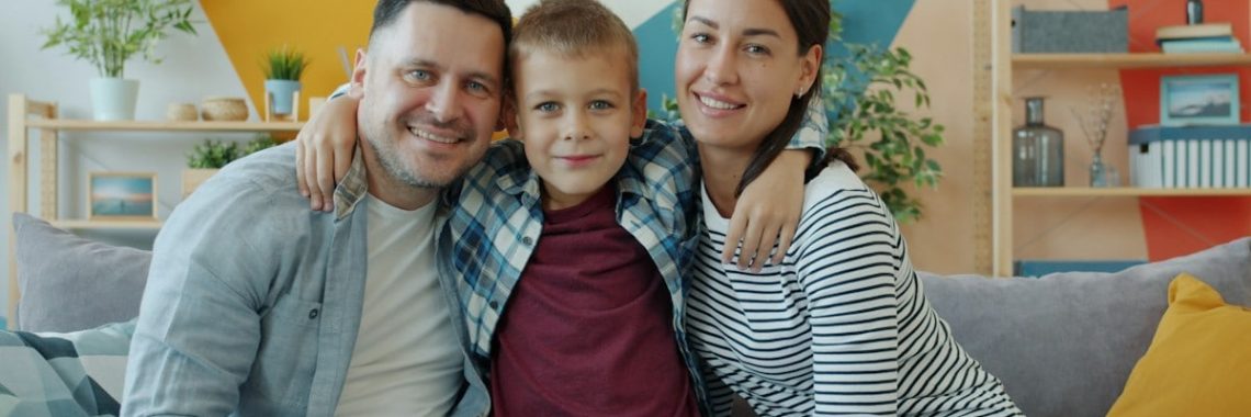 portrait of happy family mother, father and son smiling looking at camera sitting on couch in modern apartment. People and lifestyle concept.